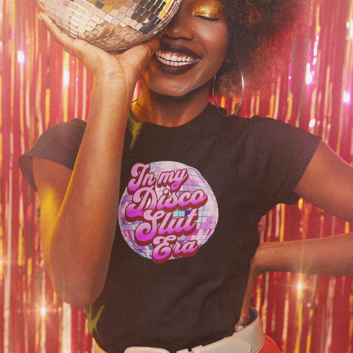 A woman with curly hair is smiling and holding a disco ball in front of her face, wearing a black t-shirt with a pink and purple design.
