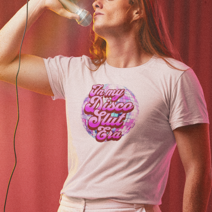 A woman wearing a white t-shirt with a graphic design of a disco ball and the text "My disco suit era" is singing into a microphone on a stage.